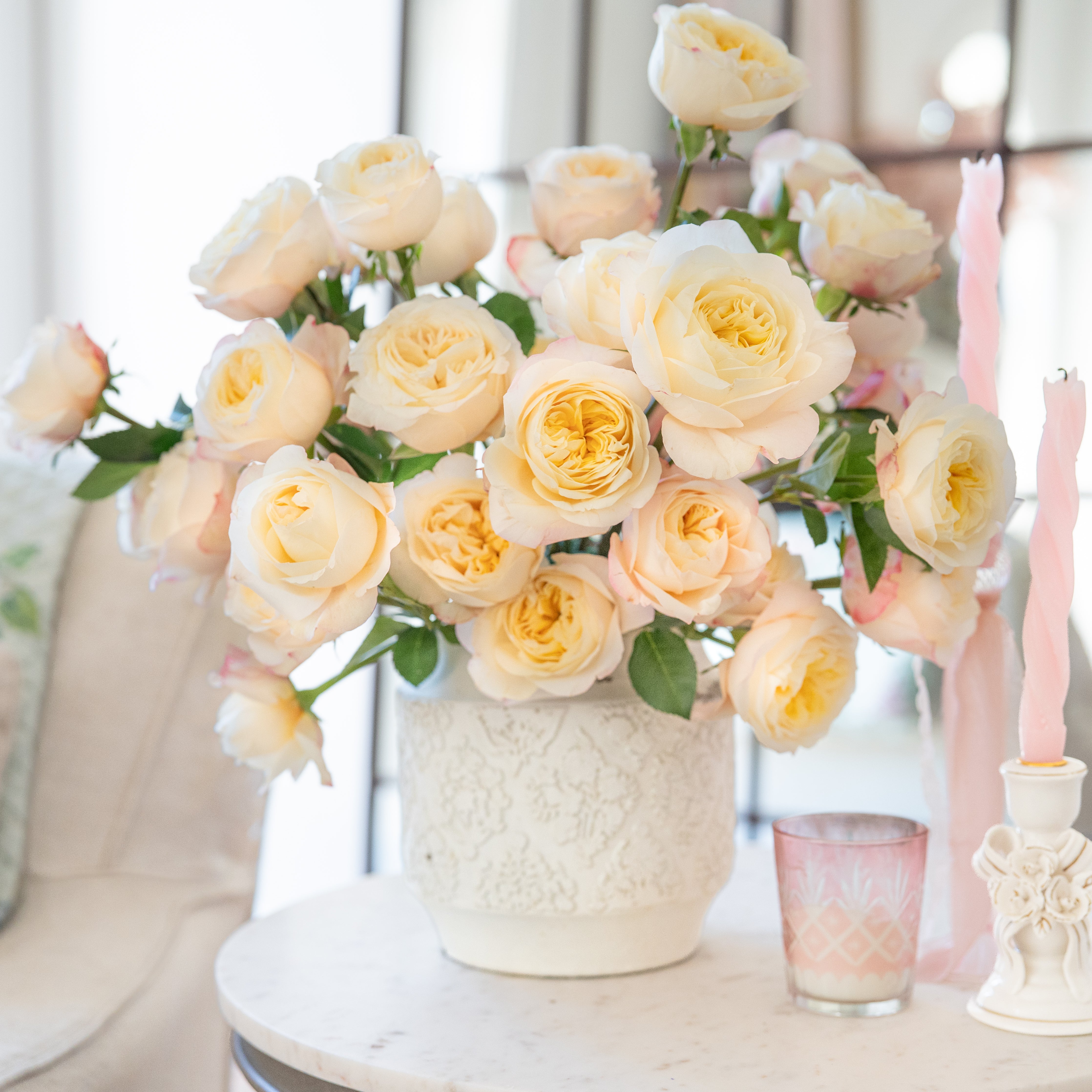Bouquet of light yellow roses in a white vase on a table with candles and a glass.