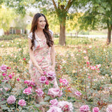 Woman in a floral dress standing among pink roses in a garden.