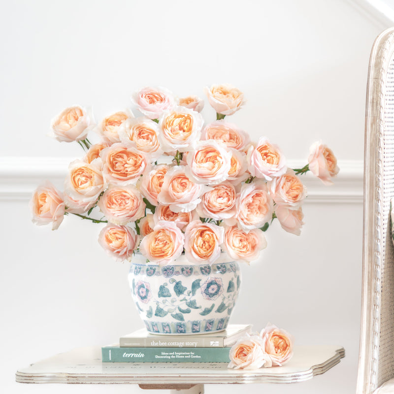 Bouquet of peach roses in a decorative vase on a table next to a wicker chair with floral cushions.
