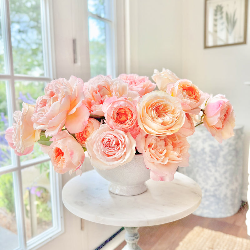 Bouquet of pink roses in a white vase on a small round table by a window.