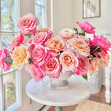 Bouquet of pink and peach roses in a white vase on a table by a window.