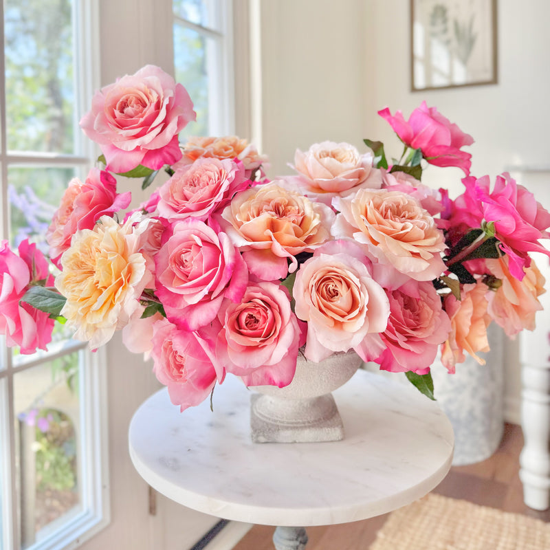 Bouquet of pink and peach roses in a white vase on a table by a window.