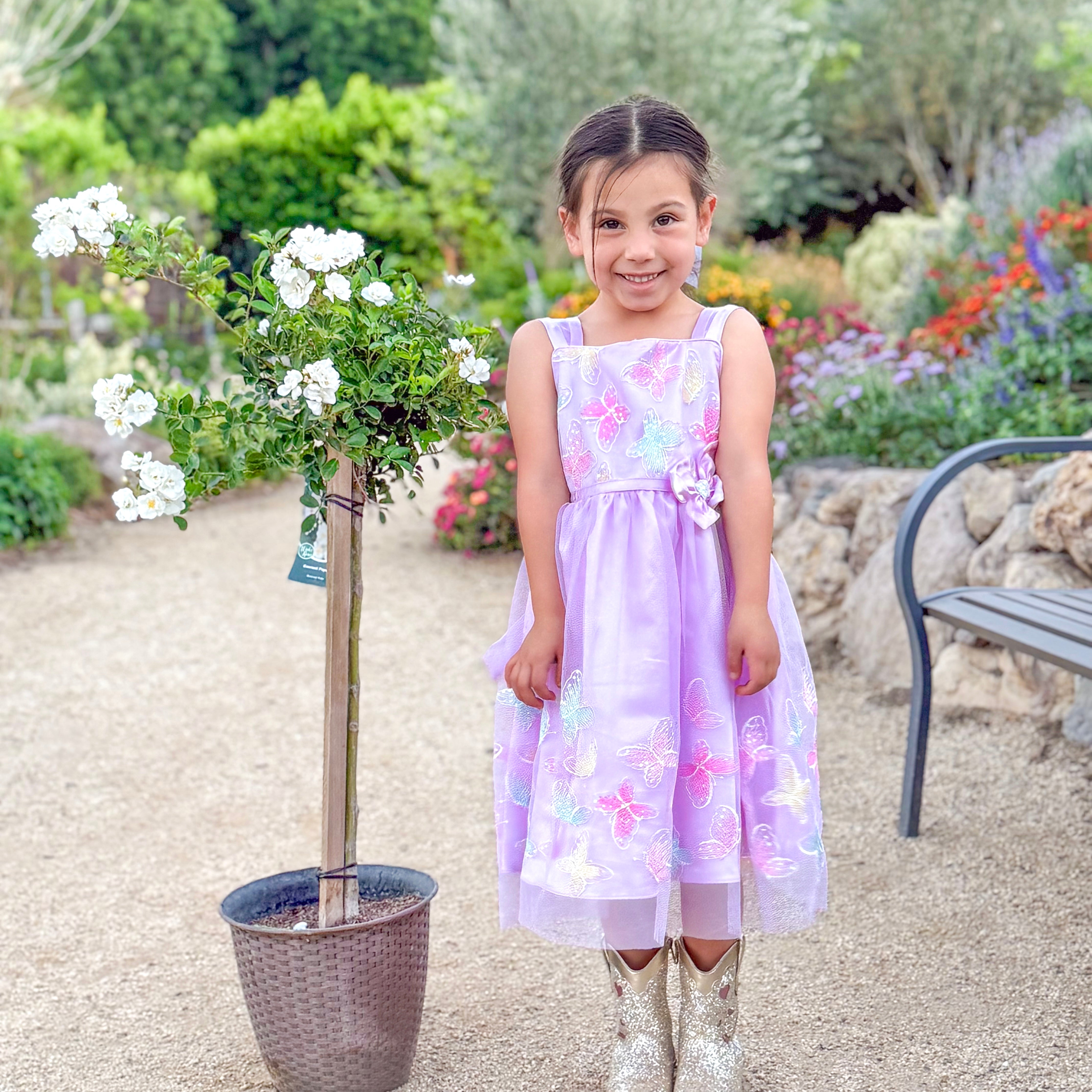 Young girl in a floral dress standing in a garden with flowers and a bench in the background.